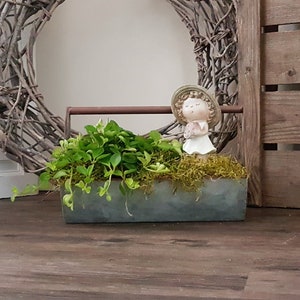 Könnte beinhalten: Ein rechteckiger Metallpflanzer gefüllt mit grünen Pflanzen und Moos, mit einer kleinen Figur eines Mädchens mit Hut. Der Pflanzer steht auf einer Holzoberfläche, mit einem geflochtenen Kranz und Holzpaneelen im Hintergrund.