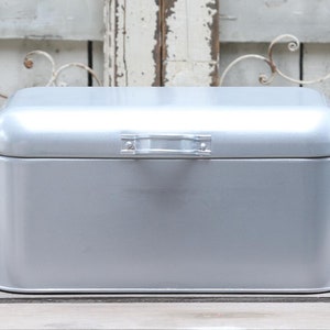 Könnte beinhalten: Eine silberfarbene Brotbox aus Metall mit gewölbtem Deckel und kleinem Griff. Die rechteckige Box hat einen Klappdeckel und ist zur Aufbewahrung von Brot und Backwaren bestimmt. Die Brotbox steht auf einer Holzoberfläche.