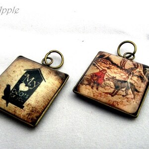 Könnte beinhalten: Zwei quadratische Anhänger mit bronzefarbenen Metallrahmen. Ein Anhänger zeigt ein Vogelhausdesign mit dem Text "My heart beats for you". Der andere Anhänger zeigt eine Szene mit einer Person in Rot und Wölfen.
