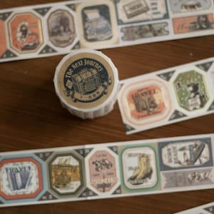 May include: A roll of decorative washi tape with a travel theme, featuring a treasure chest design and the words "The Next Journey." Several strips of tape with vintage-style travel-themed illustrations are also visible.