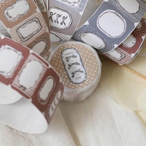 May include: A selection of washi tape rolls in shades of brown, grey, and red. The tape is decorated with label designs and bow illustrations, with the word "FRENCH" visible on one roll. Suitable for scrapbooking and craft projects.