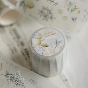 May include: A roll of decorative washi tape with a lemon and floral design. The tape features illustrations of lemons, leaves, and flowers in soft yellow, green, and white hues. The tape roll has the text "Journey in Golden Light".
