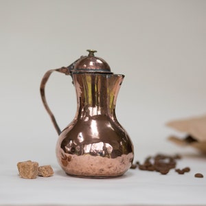 May include: A copper coffee pot with a lid and a handle. The pot is sitting on a white surface with brown sugar cubes and coffee beans.