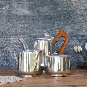 May include: A silver-toned metal tea set comprising a teapot with a curved wooden handle, a creamer, and a sugar bowl. The items feature a polished, reflective finish and are arranged on a wooden surface alongside two white artificial roses and lace.
