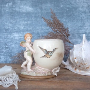 May include: A cream-coloured ceramic vase in the shape of an egg, decorated with a painted owl and a cherub figurine. A large white seashell and lace are also present. The vase is likely vintage.