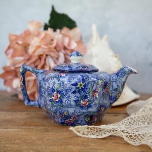 May include: A blue and white floral patterned teapot with a lid and a spout. The teapot is sitting on a wooden surface with a white lace doily and a pink flower.
