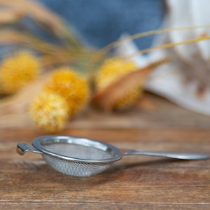 May include: A small, stainless steel tea strainer with a handle rests on a wooden surface. The strainer is round with a fine mesh. In the background, blurred yellow flowers and a white object are visible.