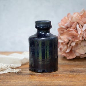May include: A dark blue ceramic bottle with a cylindrical body and a narrow neck. The bottle has a glossy finish and sits on a wooden surface. A bouquet of pink flowers and a white lace cloth are in the background.