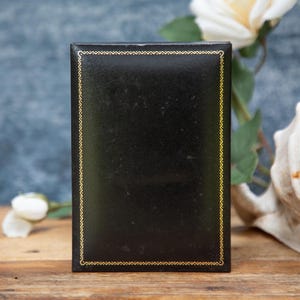 May include: A black rectangular box with a gold trim. The box is set against a backdrop of a wooden surface, a seashell, and a blue background. A white rose is also visible.