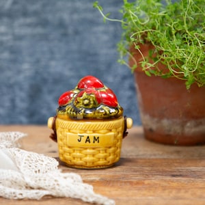 May include: A yellow ceramic jam jar with a basket weave design and the word "JAM" on the front. The lid is topped with red fruit. A potted plant and lace cloth are in the background.