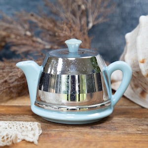 May include: A small, vintage teapot with a light blue base and handle, and a shiny, silver-colored body. The teapot is sitting on a wooden surface, with a seashell and dried plants in the background.