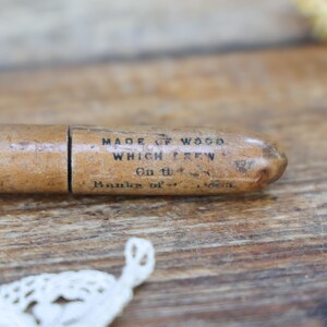 May include: A vintage wooden pencil holder with text that reads "MADE OF WOOD WHICH GREW On the Banks of..." The pencil holder is light brown and has a cylindrical shape. The background is a wooden surface.