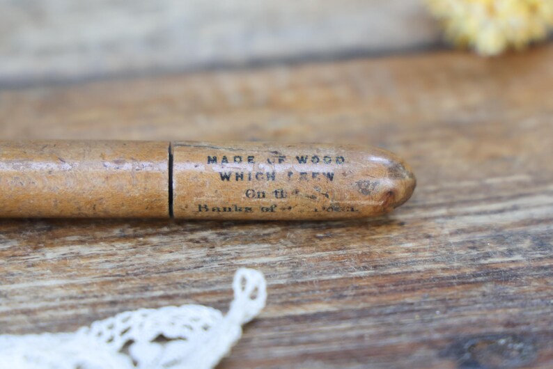 May include: A vintage wooden pencil holder with text that reads "MADE OF WOOD WHICH GREW On the Banks of..." The pencil holder is light brown and has a cylindrical shape. The background is a wooden surface.