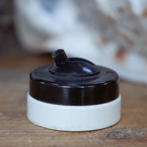 May include: Close-up of a vintage light switch. The switch has a dark brown top and a white base. The toggle is also dark brown. The switch is set against a blurred background on a wooden surface.