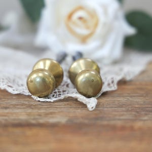 May include: Two polished brass cabinet knobs with a smooth, spherical head design. Each knob features a rounded top and a tapered base that connects to a metal screw post, showcasing a metallic gold finish with subtle reflective highlights.