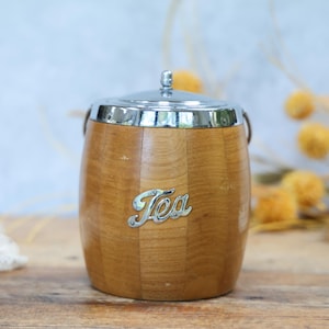 May include: A wooden tea canister with a silver lid and handle. The canister is barrel-shaped with a light brown finish and the word "Tea" in silver script on the front. The canister sits on a wooden surface.