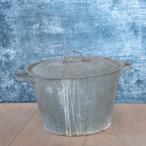 May include: A vintage metal pot with a lid, showing signs of age. The pot is made of galvanized metal, with side handles and a loop on the lid. White streaks mark the exterior. It sits on a wooden surface, against a blue background.