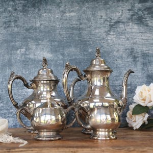 May include: Two antique silver tea sets with ornate handles and lids are displayed on a wooden surface. The tea sets are highly reflective, with a small creamer and a teapot. White roses and a lace cloth are also present.