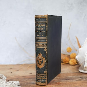 May include: An antique book titled "Life & Times of Queen Victoria, Vol. 2." The book has a black cover and a spine with gold lettering and decorative elements. The book is standing upright on a wooden surface.