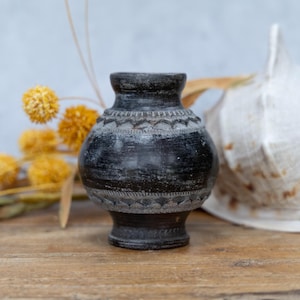 Könnte beinhalten: Eine kleine, dunkelgraue Vase mit geätzten Mustern am Körper und am Hals. Die Vase steht auf einer Holzoberfläche, mit getrockneten gelben Blumen und einer großen Muschel im Hintergrund. Die Vase hat einen runden Körper und einen schmalen Hals.