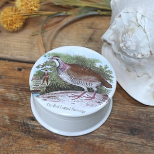 Può includere: Scatola portagioie rotonda bianca con coperchio con illustrazione di una Pernice rossa. La scatola è su una superficie di legno, con fiori secchi e una conchiglia sullo sfondo. Il testo "The Red Legged Partridge" è visibile.
