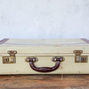 Può includere: Valigia beige vintage con finiture bordeaux e manico marrone scuro. La valigia ha chiusure color ottone e una serratura. L'esterno è strutturato e si trova su una superficie di legno.