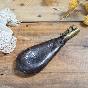 May include: A dark brown, textured leather powder flask with a brass nozzle and a mechanical spring-loaded dispensing mechanism. The flask has a teardrop shape and a small metal eyelet near the neck, positioned next to white lace fabric.