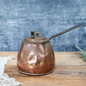 May include: A hammered copper pot with a rounded body, a flat lid featuring a small rectangular handle, and a long, straight metal handle extending from the side. The surface of the copper shows visible indentations and a warm, metallic finish.