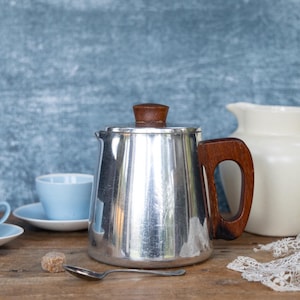 May include: A stainless steel coffee pot with a wooden handle and lid, alongside light blue teacups and saucers, a white pitcher, and a spoon. The items are arranged on a wooden surface.