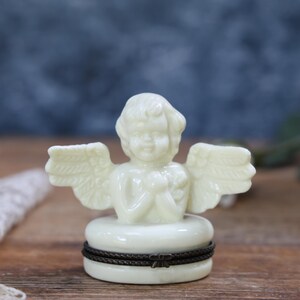 May include: A small, off-white, opaque figurine of a cherub with wings, positioned atop a circular base. The base features a dark, textured, horizontal band wrapped around its circumference, creating a distinct contrast with the light-colored material.