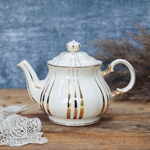 May include: White ceramic teapot with gold accents and vertical stripes. The teapot has a lid with a crown-shaped finial and a curved handle. The design is elegant and vintage, suitable for serving tea.