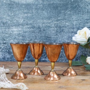 May include: A set of four copper-toned goblets with metallic stems and circular bases. Each goblet features a smooth, reflective finish and a flared bowl shape. The set is arranged in a row, showcasing a consistent design across all four pieces.