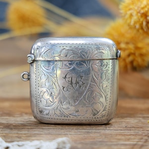 May include: A silver-toned metal match safe featuring an intricate engraved floral pattern across the front surface. The rectangular container has rounded corners, a hinged lid, and a small circular loop attached to the side for hanging.