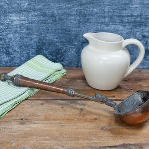 May include: A copper and brass ladle with a wooden handle rests on a wooden surface next to a white ceramic jug and a green and white checkered cloth. The ladle has an ornate handle.