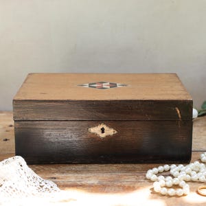 May include: A rectangular dark brown wooden box with a hinged lid featuring a central diamond-shaped inlay pattern of red, black, and white. The front of the box displays a brass keyhole escutcheon plate centred on the dark wood grain surface.