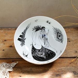 May include: A white ceramic bowl featuring a black ink-style illustration of two figures in flowing garments, a peacock feather, and a tall structure. The design includes intricate patterns and small bird motifs printed on the interior surface.