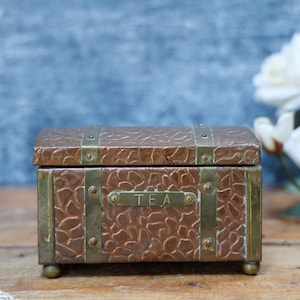 May include: A rectangular copper tea chest with a textured, pebble-like surface and a hinged lid. The box features vertical brass straps secured with rivets and a central brass plaque engraved with the word TEA. It stands on four small brass feet.
