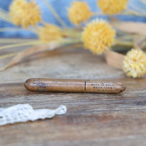 May include: A vintage wooden pencil holder with the text "MADE OF WOOD WHICH IS NY" printed on it. The pencil holder is brown and cylindrical, with a slightly worn appearance. Yellow dried flowers are in the background.