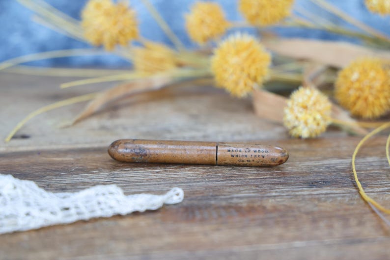 May include: A vintage wooden pencil holder with the text "MADE OF WOOD WHICH IS NY" printed on it. The pencil holder is brown and cylindrical, with a slightly worn appearance. Yellow dried flowers are in the background.