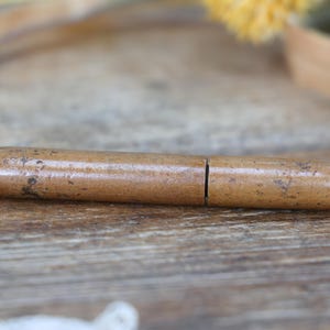 May include: A vintage, cylindrical wooden container with a light brown finish and a split in the middle. The container has rounded ends and a slightly textured surface, resting on a wooden surface.
