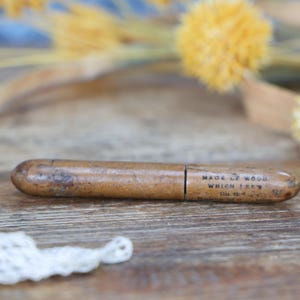 May include: A vintage wooden pencil case with a worn, brown finish. The case is cylindrical with a sliding lid and features the text "MADE OF WOOD. WHICH I'VE..." The background includes yellow flowers and a white lace trim.
