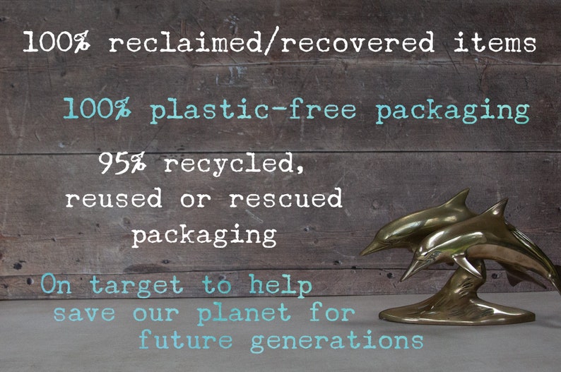 May include: A bronze dolphin sculpture with two dolphins leaping from the water. The background is a rustic wooden surface with text that reads "100% reclaimed/recovered items" and "100% plastic-free packaging."