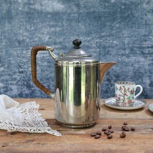Puede incluir: Una cafetera plateada con asa trenzada y pomo negro sobre una superficie de madera. Dos tazas de té florales y platillos junto a ella, con granos de café y un tapete de encaje. El fondo es una pared azul grisáceo.