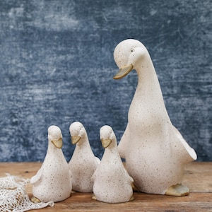 May include: A set of four off-white ceramic duck figurines with brown speckles and gold-toned beaks and feet. The collection includes one large standing duck and three smaller ducklings, all featuring a smooth, matte finish and rounded shapes.