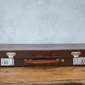 May include: A vintage brown leather briefcase with a handle and metal clasps. The rectangular case sits on a wooden surface, suggesting a classic or antique aesthetic. The briefcase is closed.