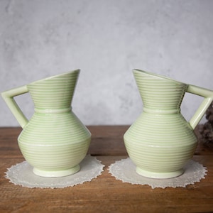 May include: Two green ceramic jugs with a striped design. The jugs have a wide base and a narrow neck. They are sitting on white doilies on a wooden surface.