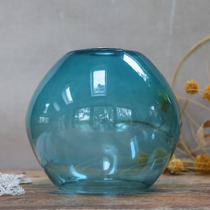 May include: A teal-coloured, spherical glass vase with a wide opening. The vase reflects the surrounding environment, including a window. Dried yellow flowers and a lace doily are in the background. The vase is on a wooden surface.