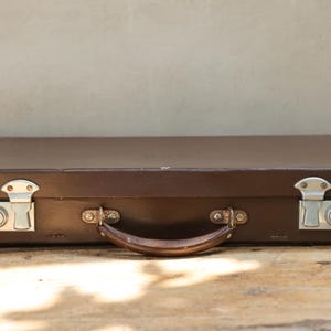 May include: A rectangular brown hard-shell briefcase featuring a central handle and two silver-toned metal locking latches on the front. The case has a smooth, solid-colored exterior finish with visible stitching along the edges and metallic hardware.