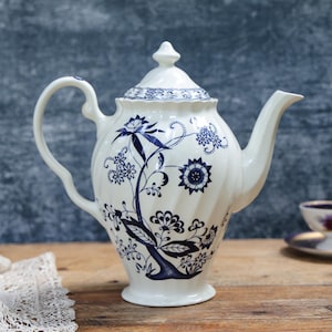 May include: A white ceramic teapot with a blue floral pattern. The teapot features a curved handle, a spout, and a lid with a finial. A matching teacup and saucer are in the background. The teapot is on a wooden surface.