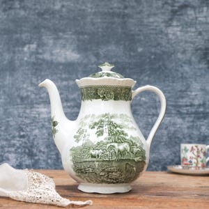May include: A white ceramic teapot with a green pastoral scene design, a matching lid, and a curved handle. Two small teacups with saucers are in the background. A white lace doily is in the foreground. The teapot is approximately 25 cm tall.